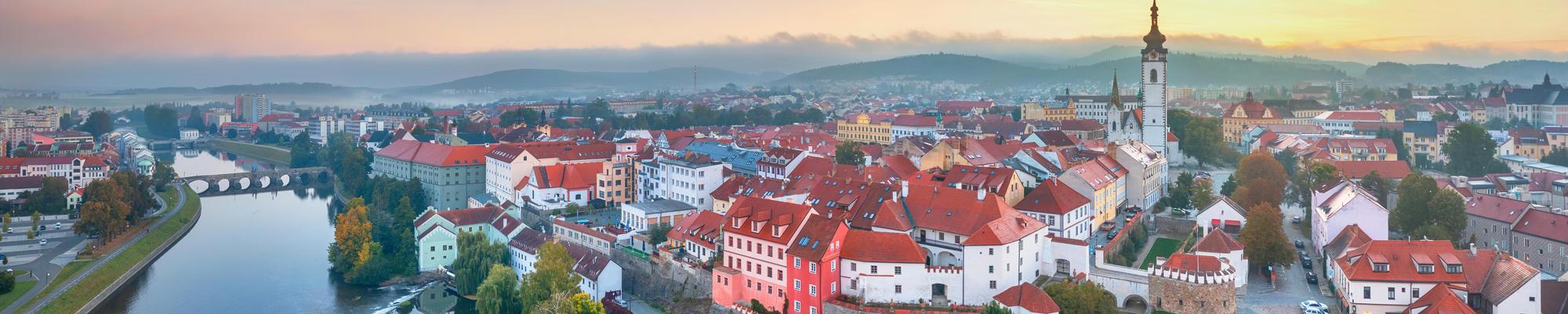 A picture of Pisek with the river and its bridges on the left side and an idyllic impression from the medium-sized town on the right. In the background small mountains are visible in what's either the beginning of sunset or end of a sunrise.