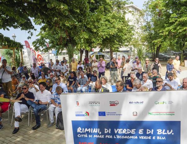 A picture of a large group of people sitting down outside listening to a presentation. In the foreground is a banner including Re-Values logo and a statement to many of the projects being funded by European funds