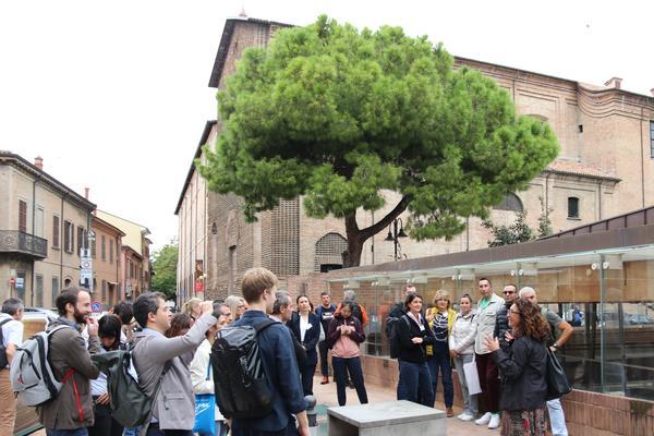 A group of people standing around listening to a presenter outside. The background includes some buildings that look like heritage buildings and a beautiful tree. 